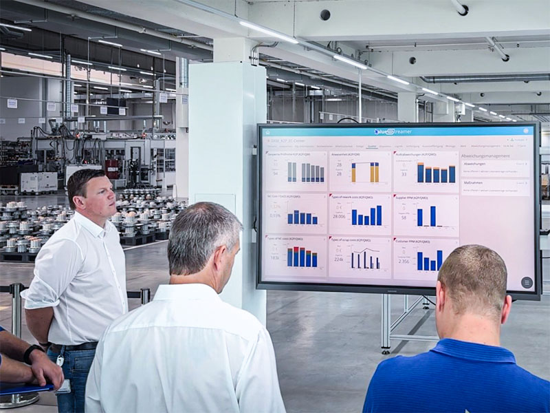 Shopfloor-Runde bei Ziehl-Abegg: drei Führungskräfte stehen in der Fertigungshalle vor einem großen Bildschirm mit dem ValueStreamer-Board ihres Bereichs.