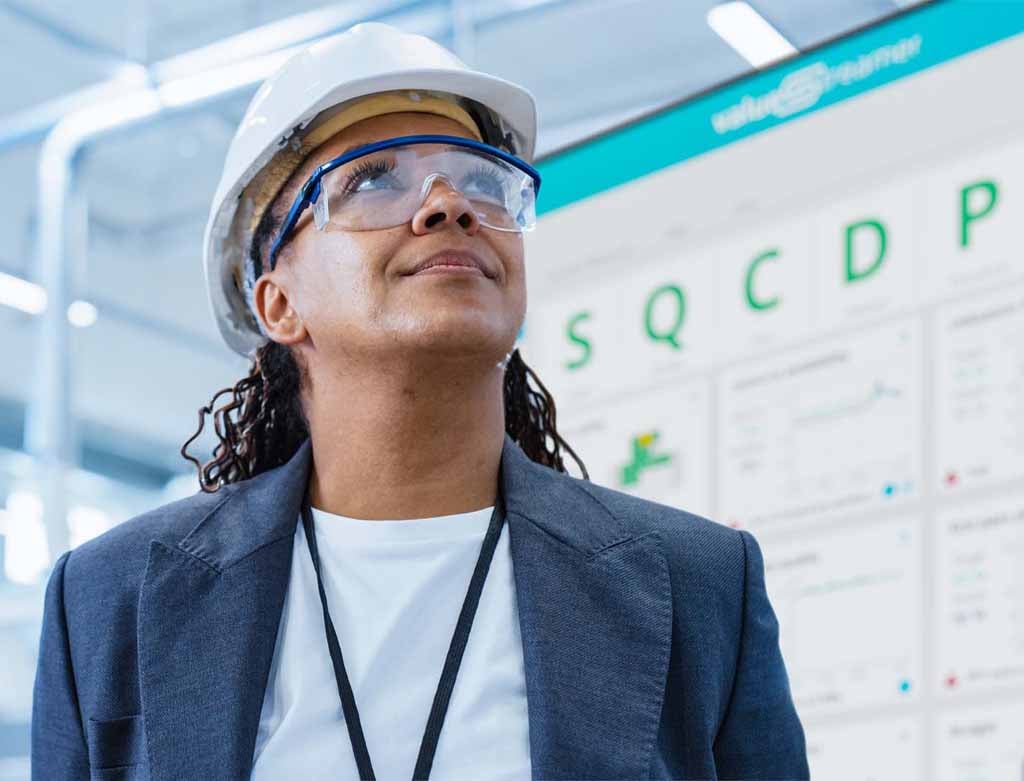 Woman wearing safety goggles and hard hat stands in front of digital Shop Floor Board from ValueStreamer