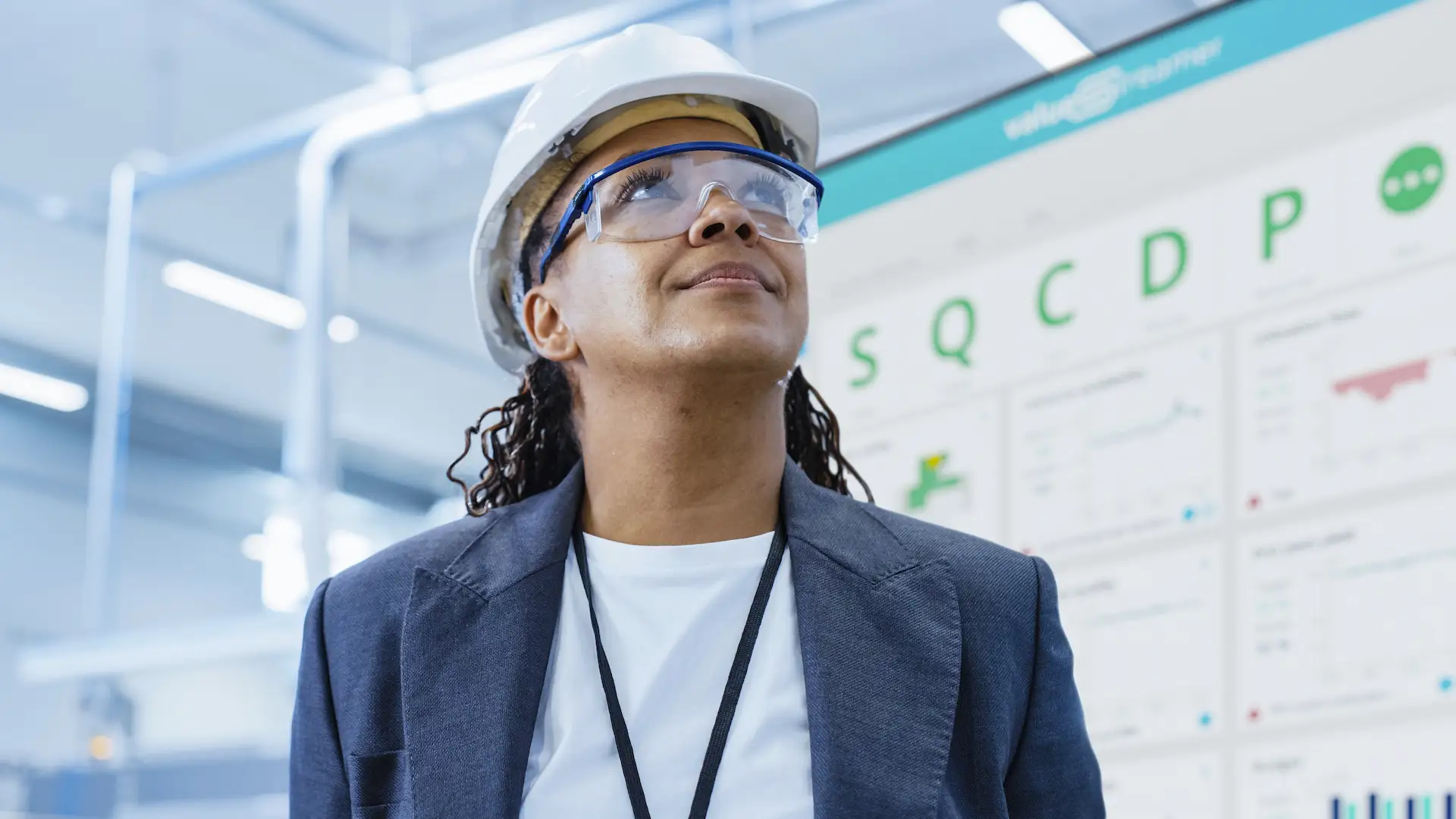 Woman wearing safety goggles and hard hat stands in front of digital Shop Floor Board from ValueStreamer